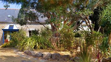 A stone building with a blue roof, surrounded by a garden with various plants and a wooden fence.