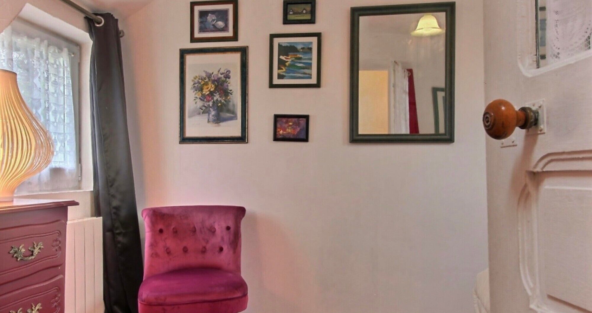 Une pièce avec une chaise rose, une commode à tiroirs, un miroir et plusieurs tableaux encadrés accrochés au mur.