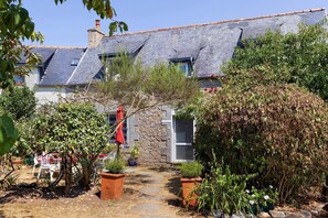 Une maison en pierre avec un toit en tuiles, entourée d’un jardin où poussent différentes espèces de plantes, et un chemin menant à l’entrée.