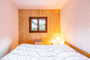A bedroom with a wooden paneled wall, a window with a view of greenery, a bed with white bedding, and a bedside lamp. - Apartment 'Appartement Résidence Ours Blanc' with Mountain View, Shared Terrace and Wi-Fi (Les Houches)