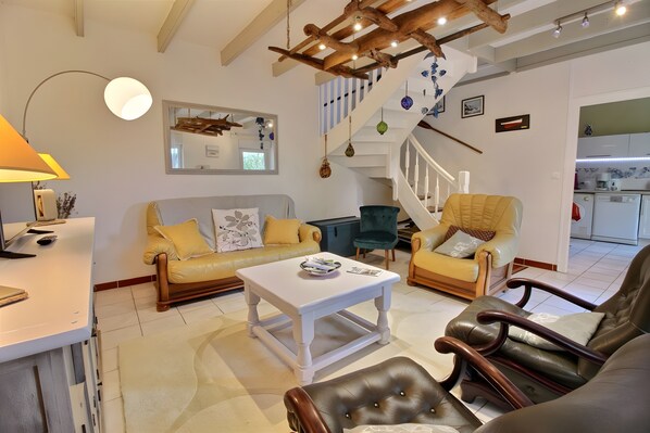 A living room with a sofa, chairs, a coffee table, a lamp, a staircase, and a kitchen in the background. - Holiday Home 'Gite Des Plages  Aigue-marine' with Private Terrace, Private Garden and Wi-Fi (Lannion)