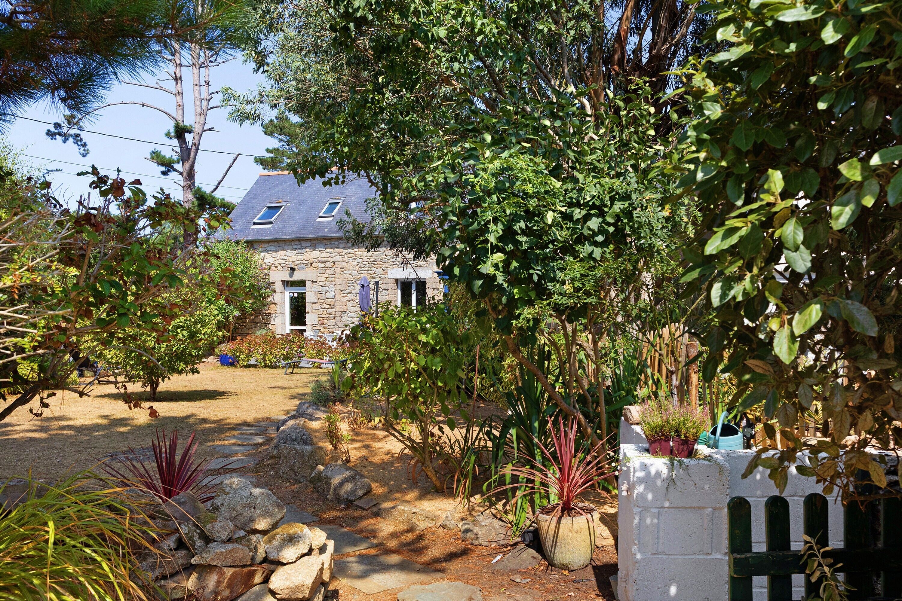 Una casa de piedra con techo inclinado, rodeada por un jardín con diversas plantas y un camino de grava que lleva a la entrada.