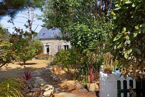 Una casa de piedra con techo inclinado, rodeada por un jardín con diversas plantas y un camino de grava que lleva a la entrada.