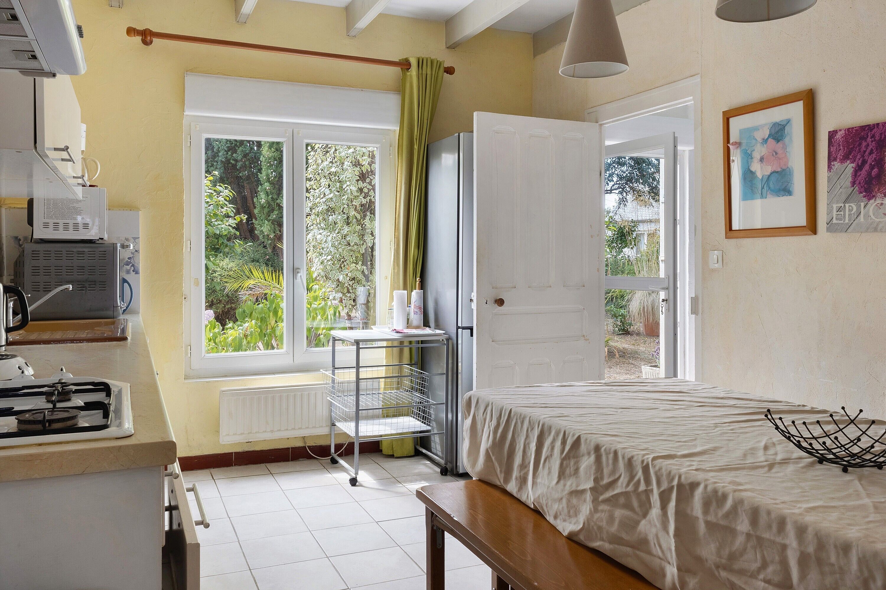 A bedroom with a bed, a window with a view of greenery, a radiator, and a small table with a lamp.