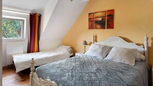 A bedroom with two single beds, a large bed with a grey quilt, a window with curtains, and a painting on the wall.