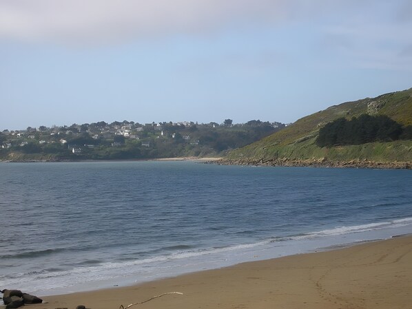 Una playa costera con arena, agua tranquila y una colina con edificios al fondo.