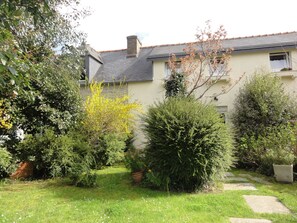 A residential garden with a pathway, various shrubs, and a house with a grey roof and beige walls.