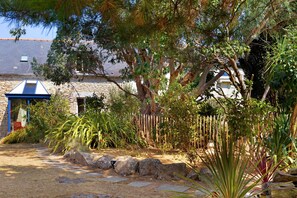 Un edificio de piedra con techo azul, rodeado por un jardín con diversas plantas y una cerca de madera.