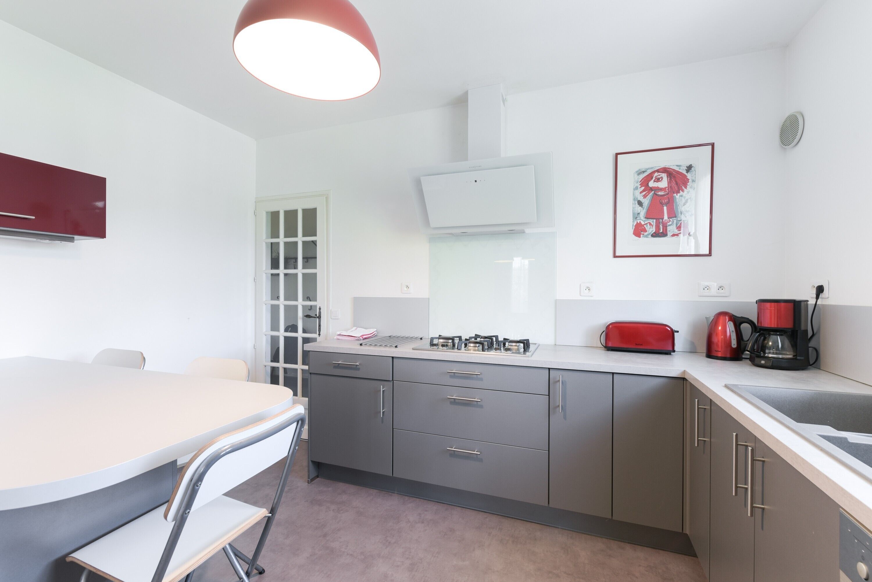 A modern kitchen with a dining table, chairs, a red toaster, a coffee maker, and a framed picture on the wall.