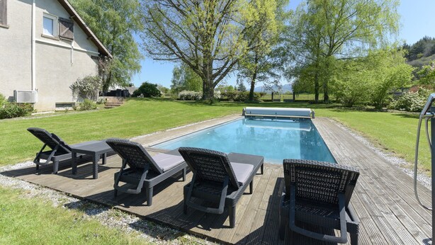 Una piscina con sillones de descanso, una terraza de madera y una casa al fondo.