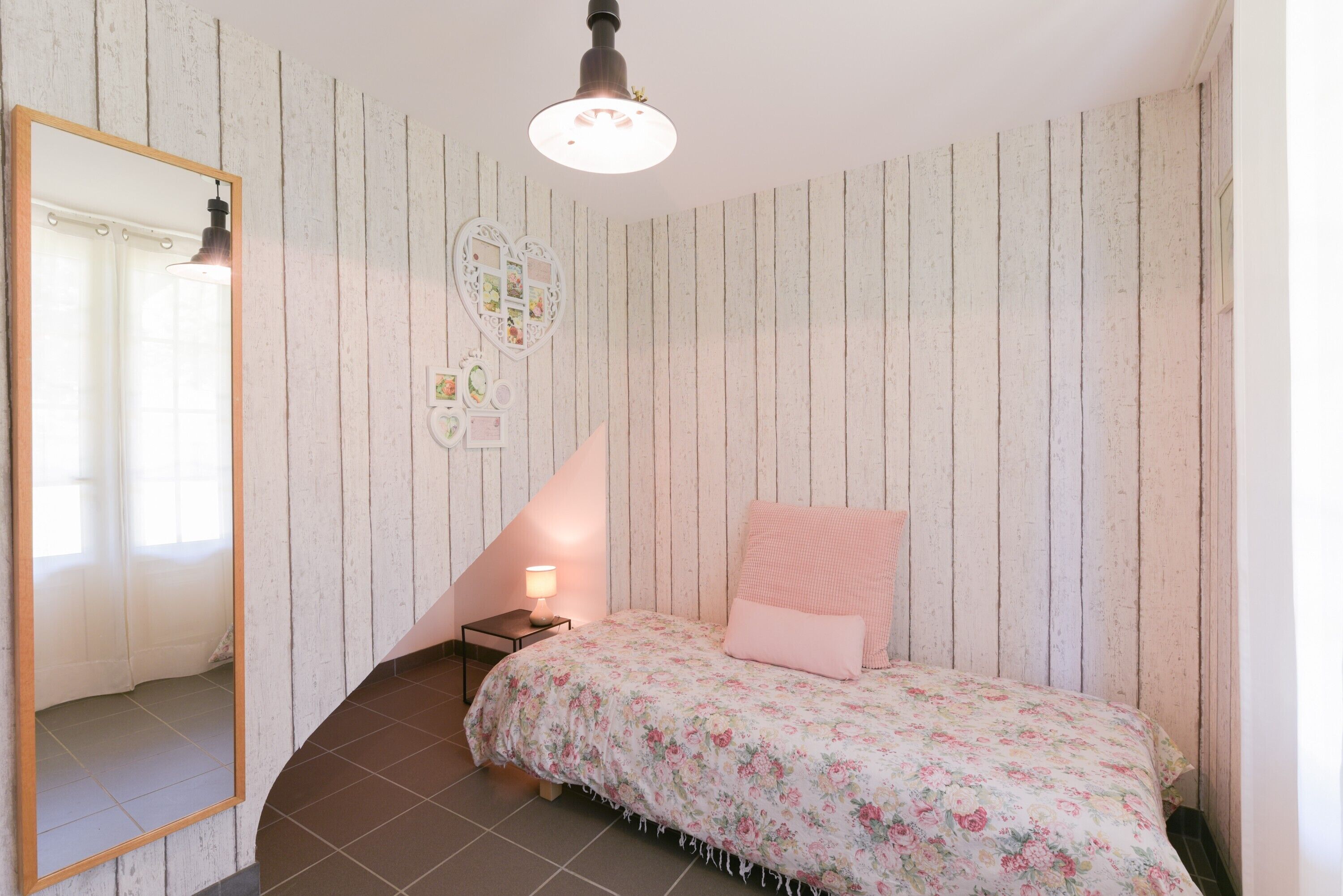 A bedroom with a wooden slat wall, a bed with floral bedding, a bedside table with a lamp, and a mirror.