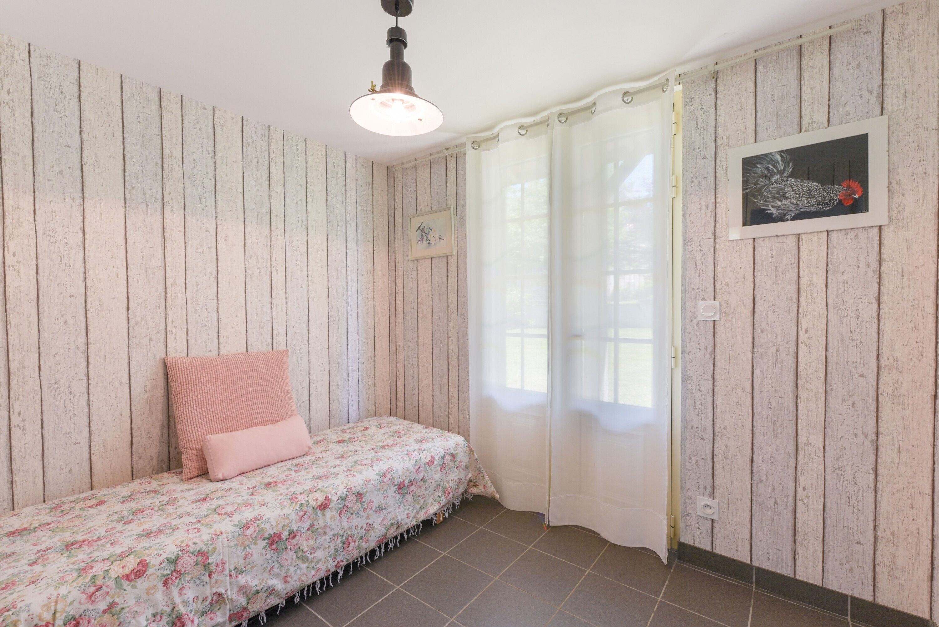 A room with wooden panel walls, a bed with floral bedding, a ceiling light fixture, a window with curtains, and a framed picture on the wall.