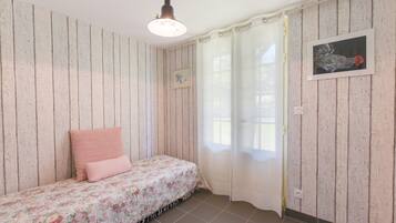 A room with wooden panel walls, a bed with floral bedding, a ceiling light fixture, a window with curtains, and a framed picture on the wall.