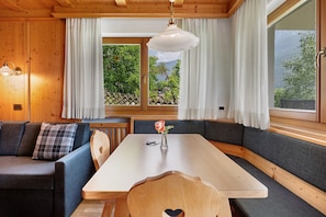 Interior de uma cabana de madeira com mesa de jantar, bancos, um sofá e uma janela com cortinas.