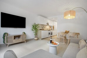 En moderne stue med et fladskærms-TV, et spiseområde med stole og et bord, et køkken med hårde hvidevarer, og en hængende lampe.