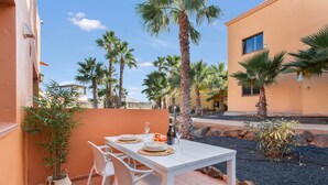 A white table with chairs and a potted plant on a tiled patio with palm trees and a building in the background.