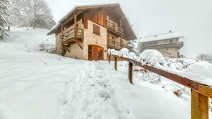 A wooden chalet with a balcony, surrounded by snow-covered trees and a ski slope.