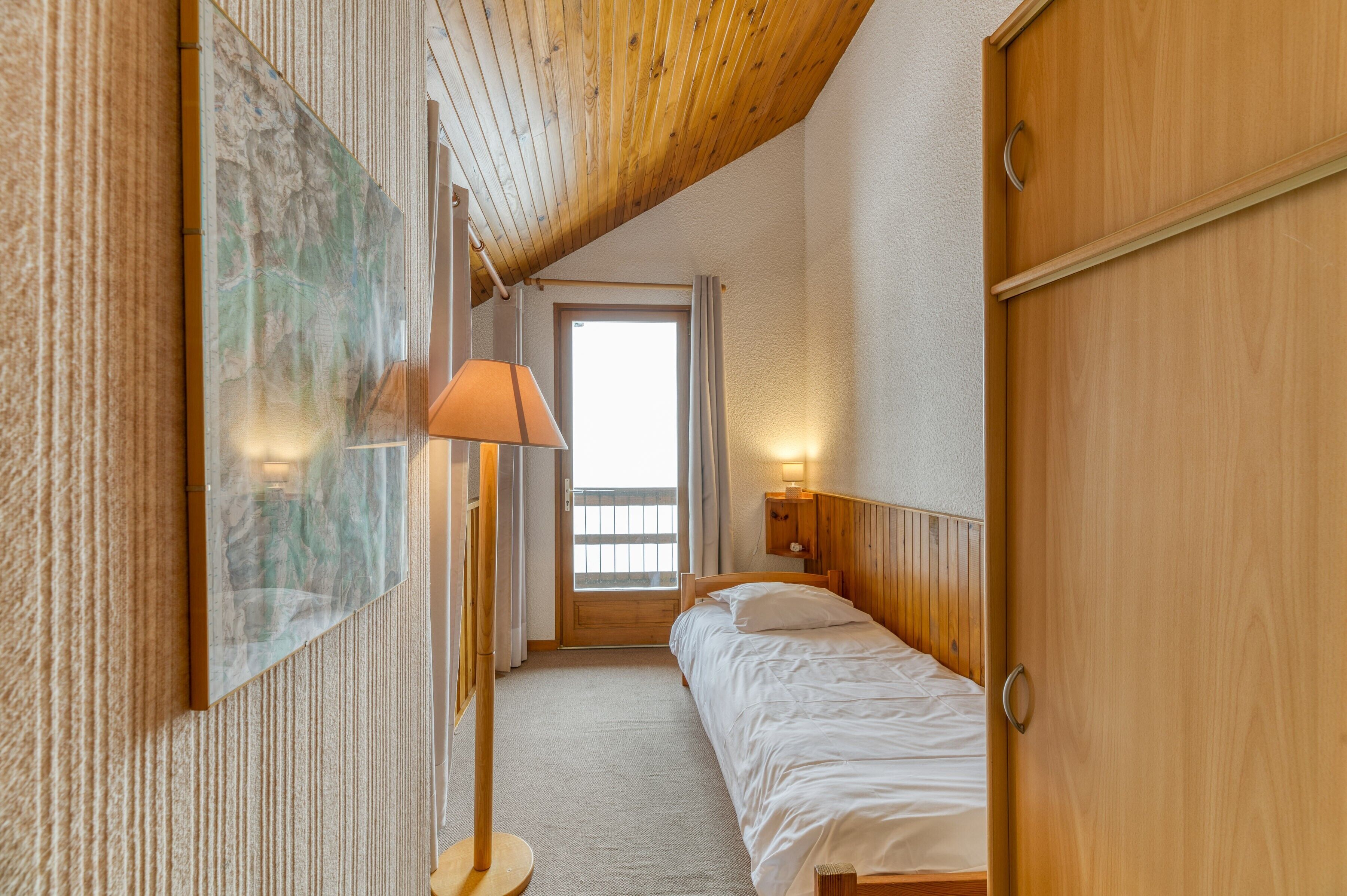 A bedroom with a wooden ceiling, a bed, a nightstand with a lamp, a wall-mounted artwork, and a door.