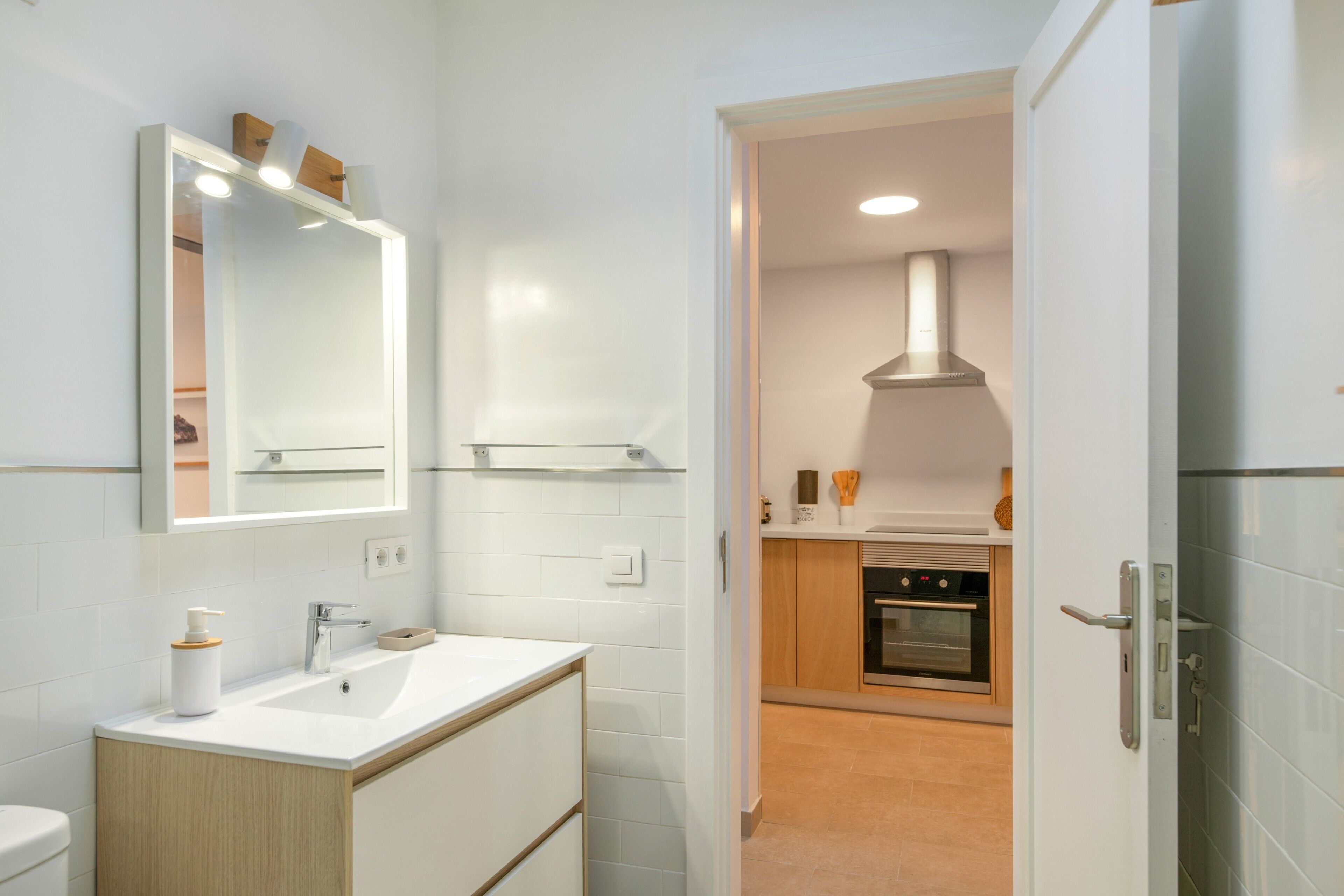 Un baño moderno con un espejo grande, un lavabo y un inodoro. Se ve un acceso a una zona de cocina con estufa y horno.