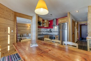 Un intérieur en bois comprenant une table à manger, des chaises, une cuisine équipée d’appareils modernes et un luminaire suspendu lumineux.