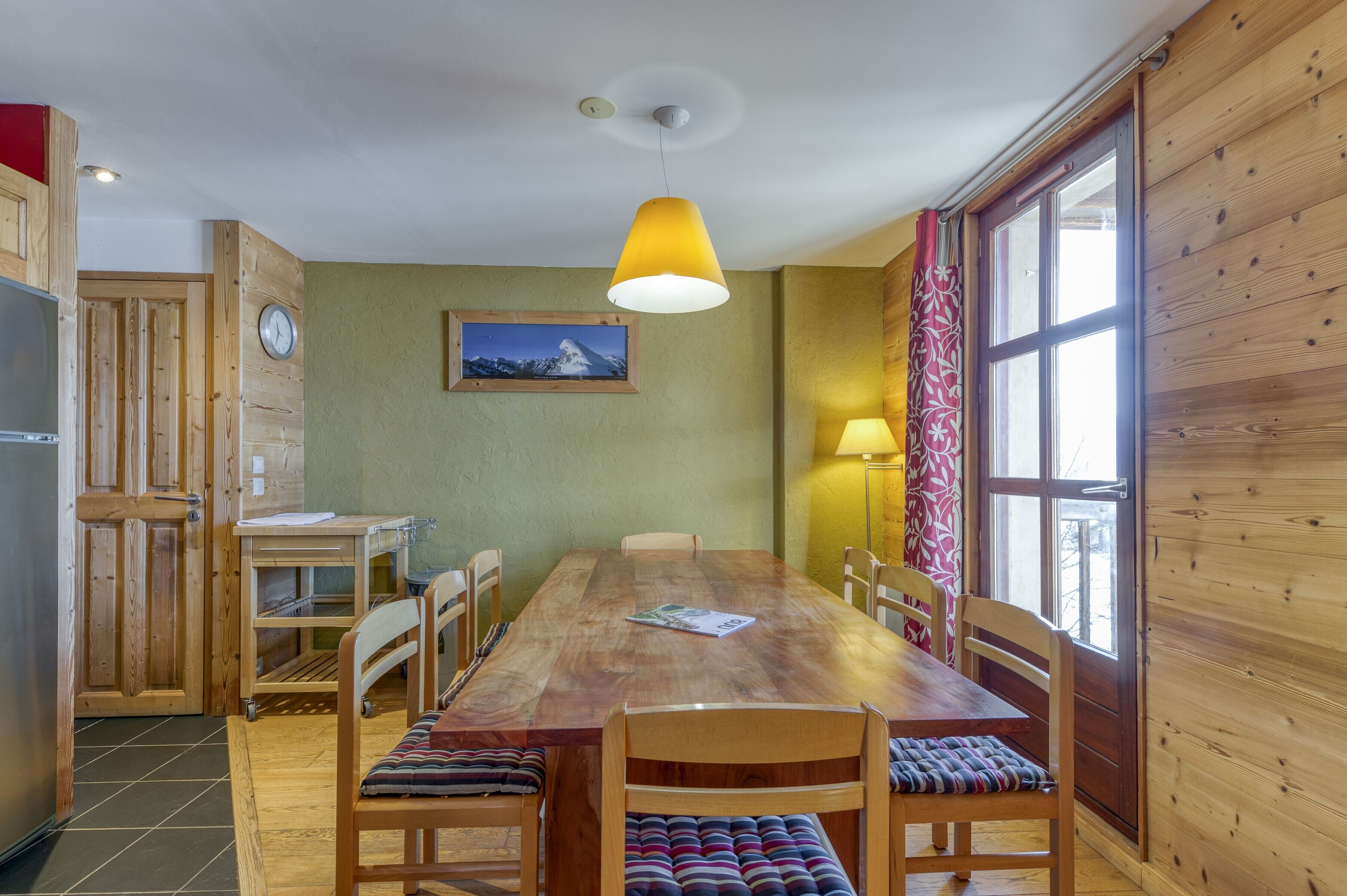 A wooden dining area with a table, chairs, a clock, a picture on the wall, and a window with curtains.