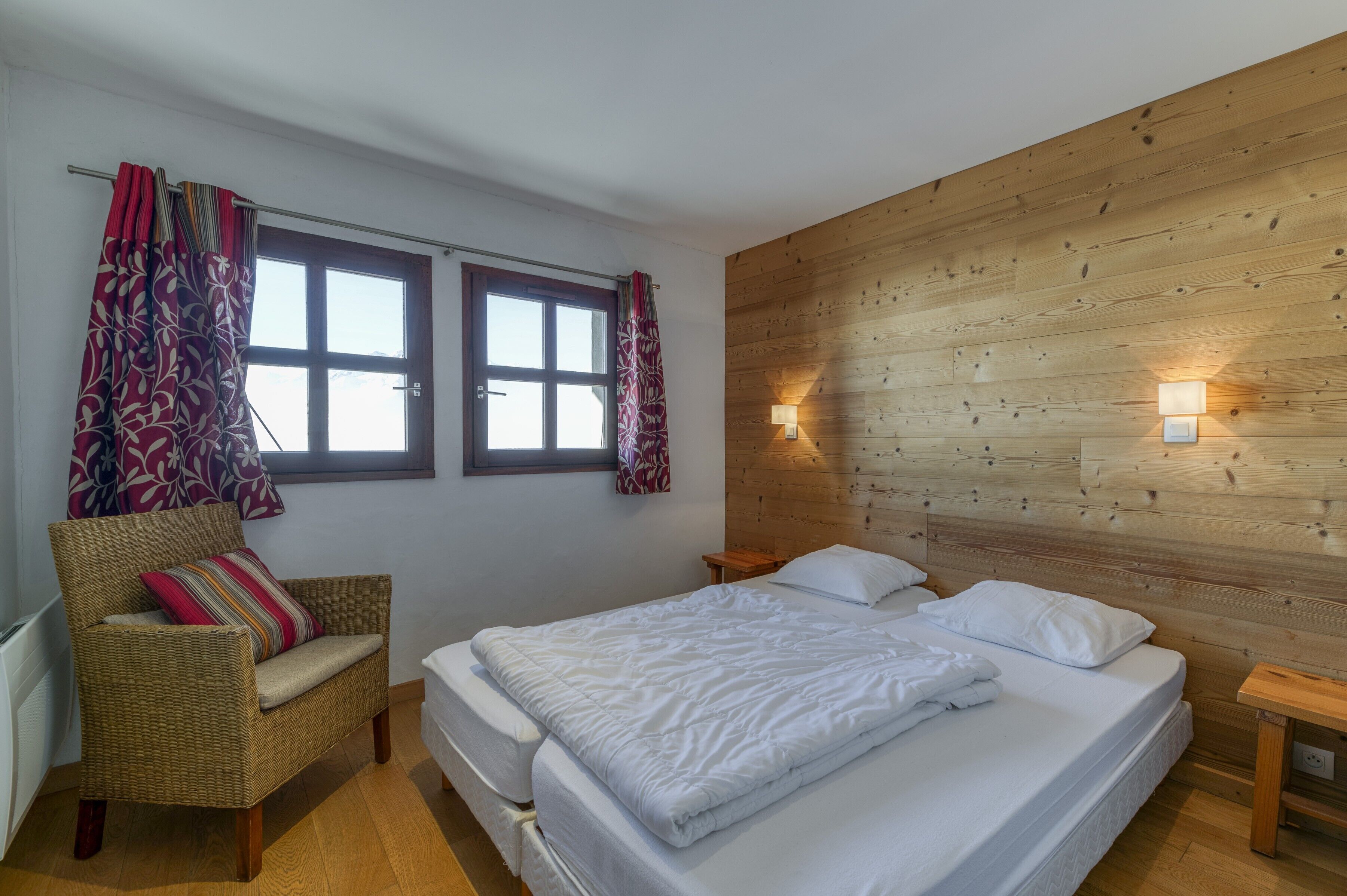 A bedroom with a wooden headboard, a bed with white bedding, a wicker chair with a striped cushion, and two windows with curtains.