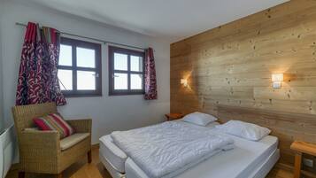 A bedroom with a wooden headboard, a bed with white bedding, a wicker chair with a striped cushion, and two windows with curtains.