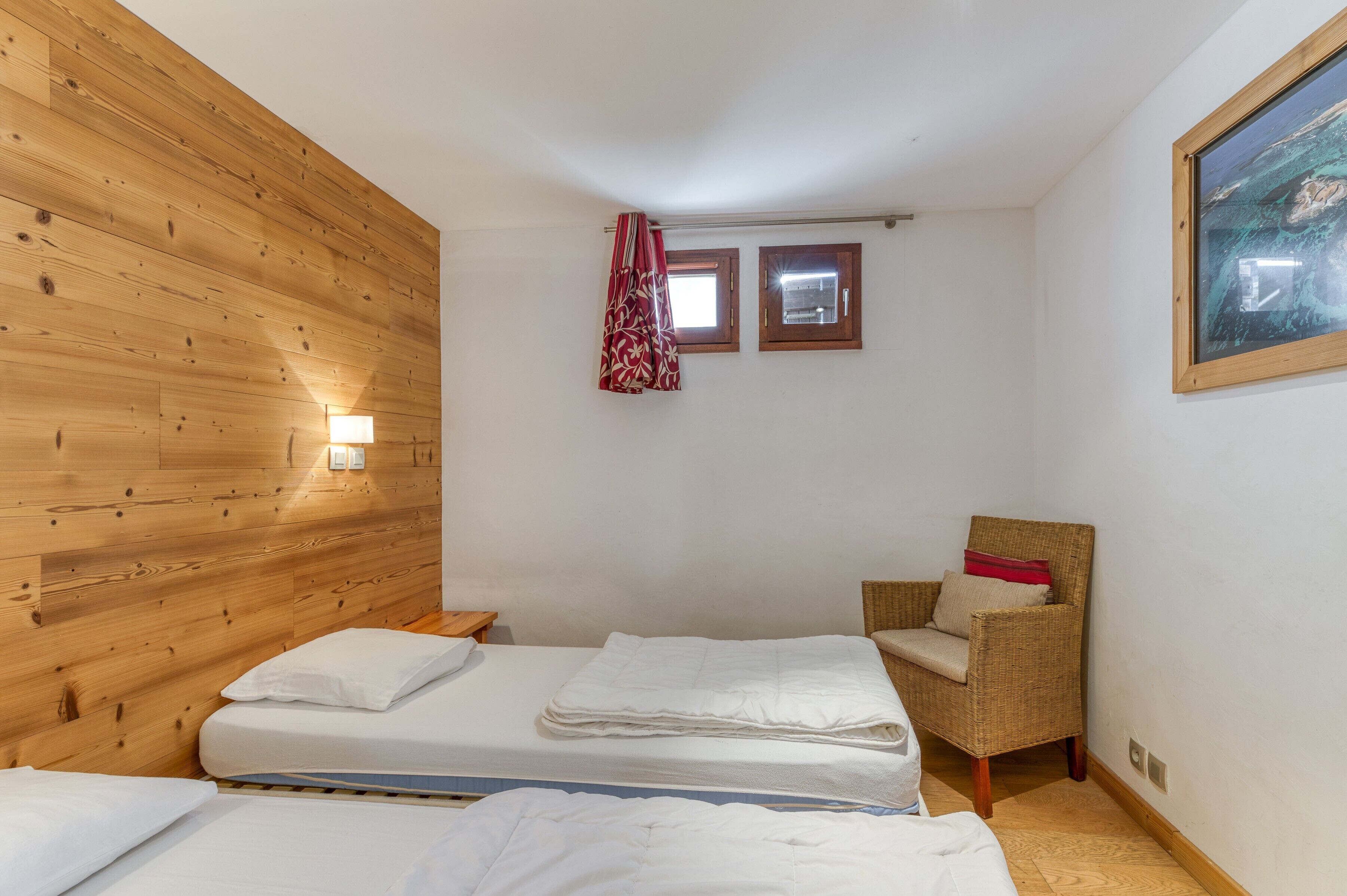 Une pièce aux murs recouverts de lambris en bois, avec deux fenêtres, un lit avec une literie blanche, une chaise en osier et un tableau accroché au mur.