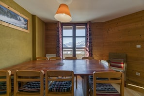 Interior em madeira com mesa de jantar, cadeiras, janela com cortinas e vista para as montanhas.