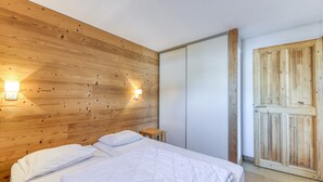 A bedroom with a wooden headboard, a bed with white bedding, a nightstand, a small table, and a wooden door. - Apartment 'Regain 1' with Mountain View and Balcony (Villard-Reculas)