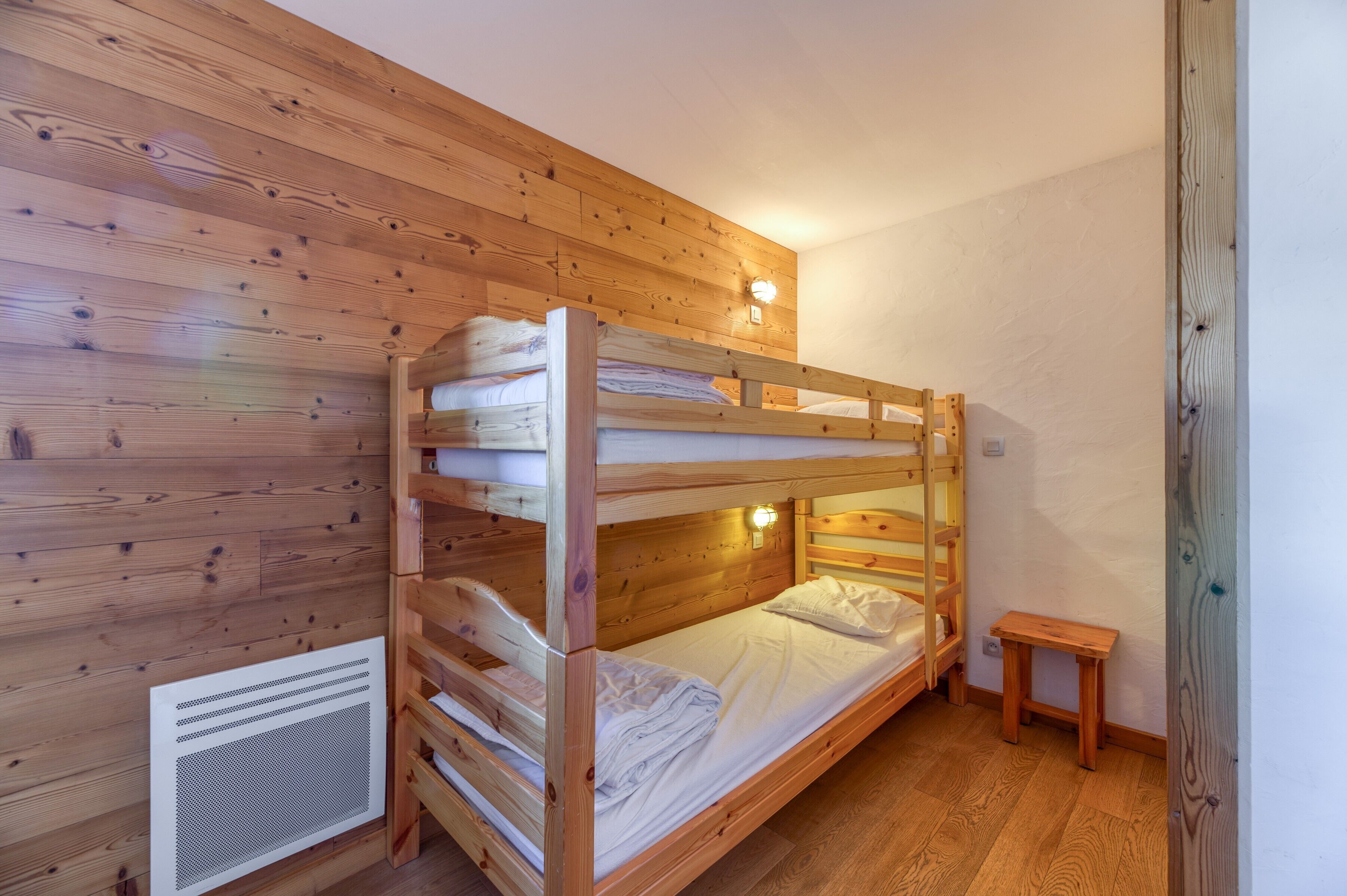 A bunk bed room with wooden walls, a small wooden stool, and a wall-mounted light.