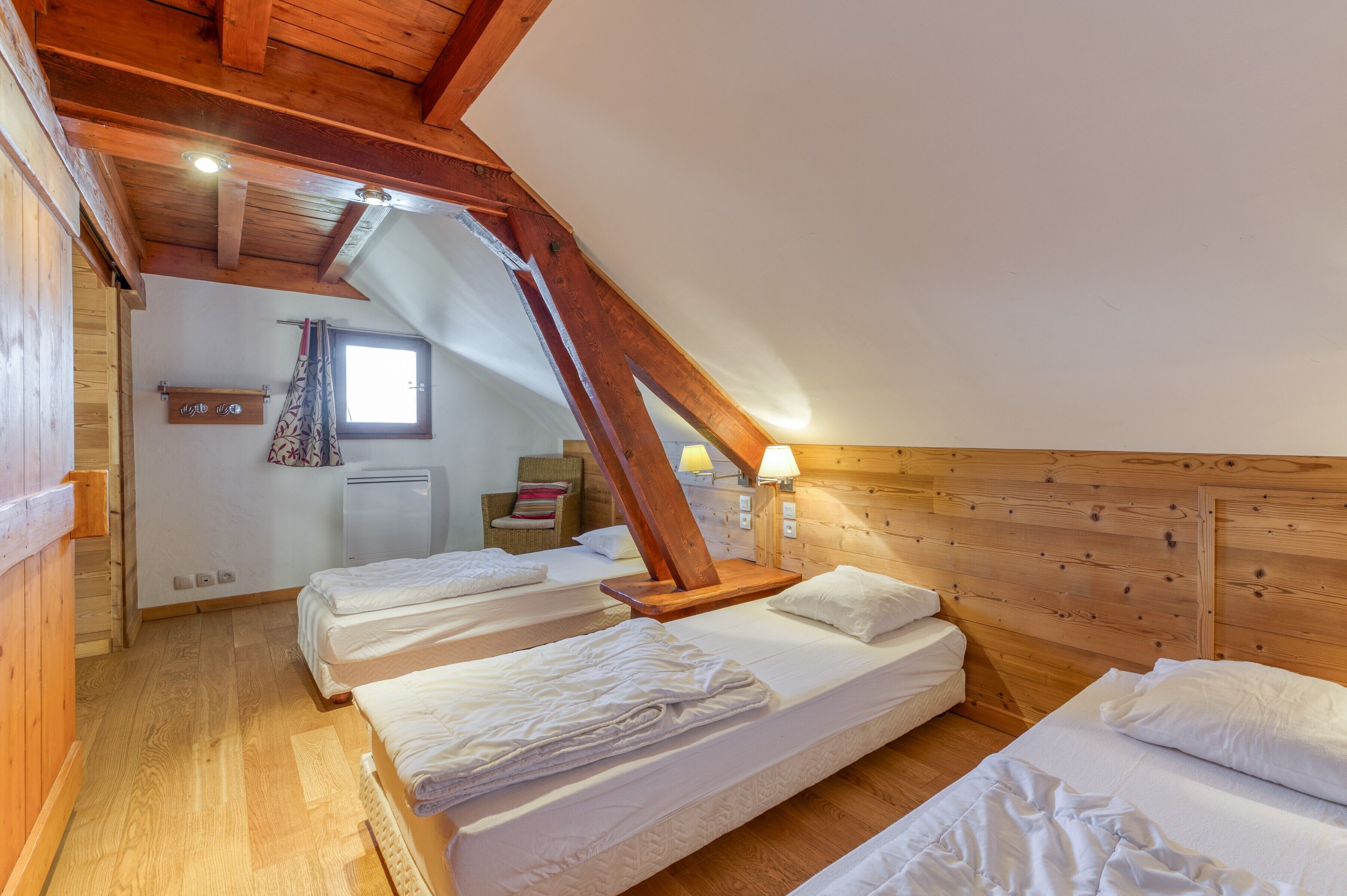 A room with three beds, wooden beams, a window with curtains, a radiator, and a small table with a lamp.