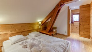 Quarto com teto de madeira inclinado, cama com roupa de cama branca, mesa de cabeceira, abajur e cadeira de madeira.