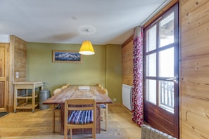 Een houten eettafel met stoelen, een geel hanglamp, een ingelijst schilderij aan de muur en een raam met gordijnen.