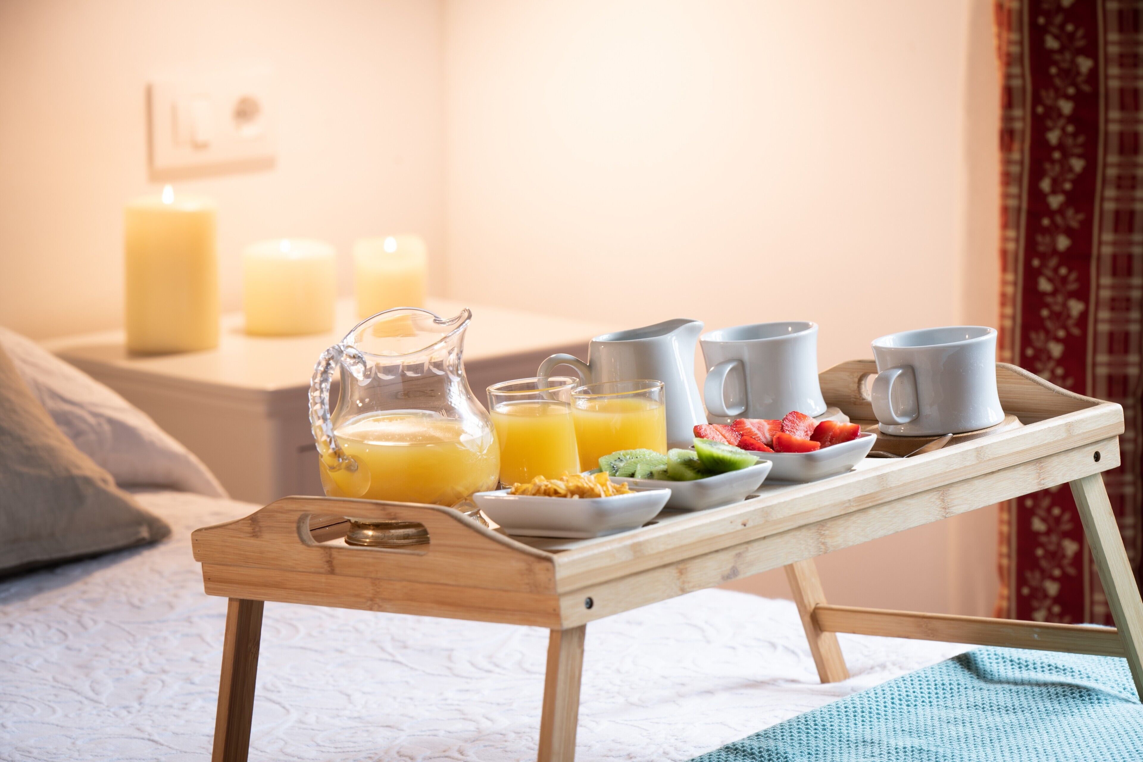 En træbakke med morgenmadsprodukter, blandt andet appelsinjuice, cereal og frugt, på en seng.