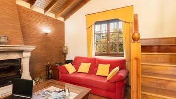 Un salón acogedor con un sofá rojo, una chimenea, un ordenador portátil sobre una mesa de centro y una ventana con cortinas.