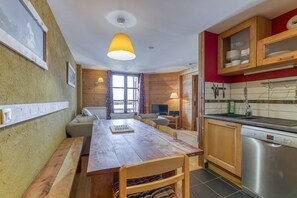 Een houten eettafel met stoelen, een houten bank, een keuken met een vaatwasser, houten kasten en een raam met gordijnen.