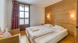 A bedroom with a wooden wall, a bed with white bedding, a wicker chair, and a window with curtains. - Apartment 'Regain 7' with Mountain View and Balcony (Villard-Reculas)