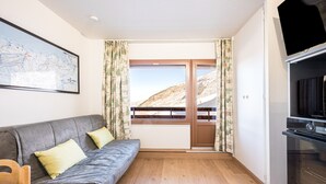 Ein Zimmer mit grauem Sofa, einem Holztisch, einem schwarzen Backofen und einem Fenster mit Blick auf eine gebirgige Landschaft.
