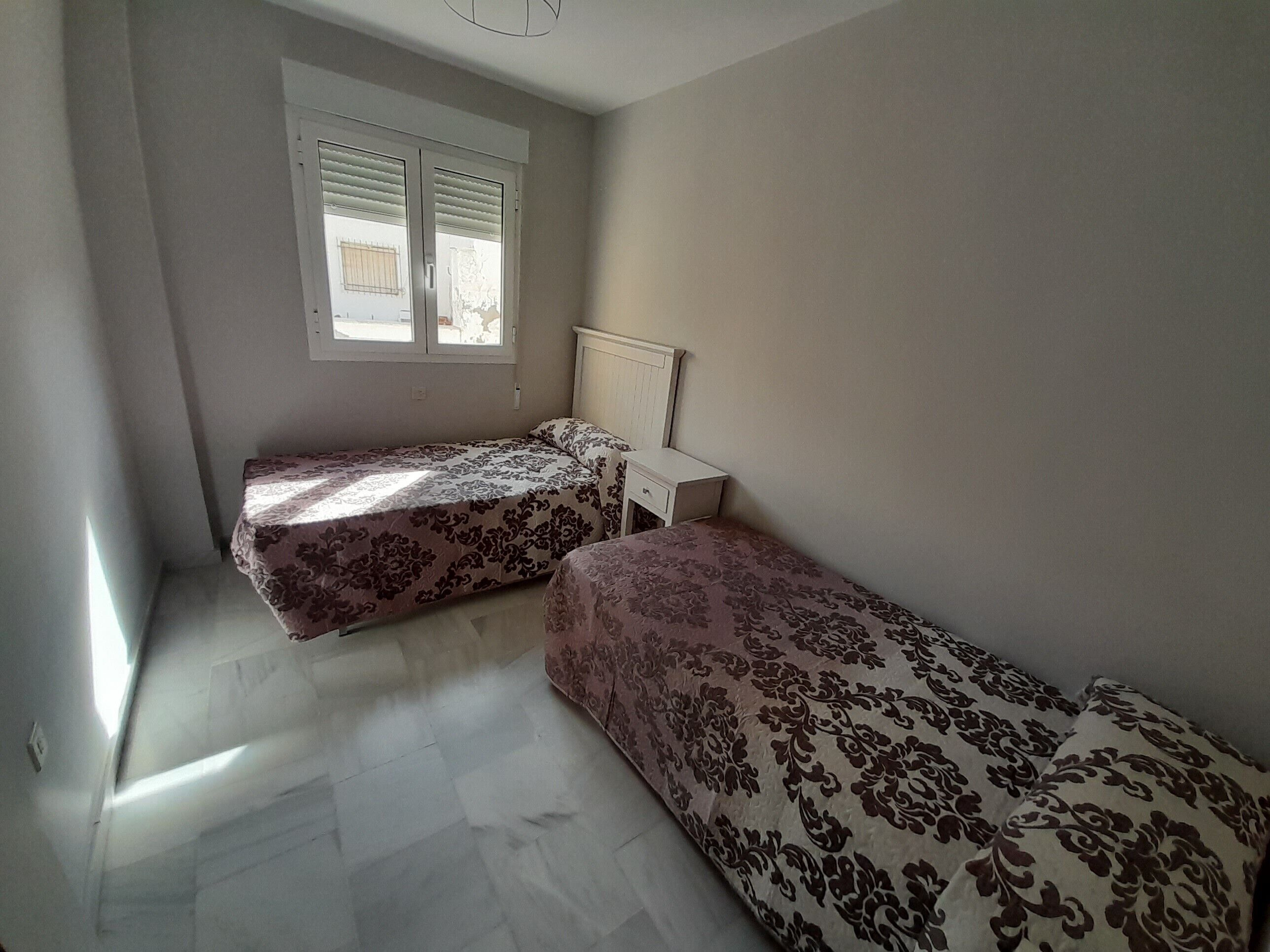 A room with two beds, a window with blinds, a white wardrobe, and a tiled floor.