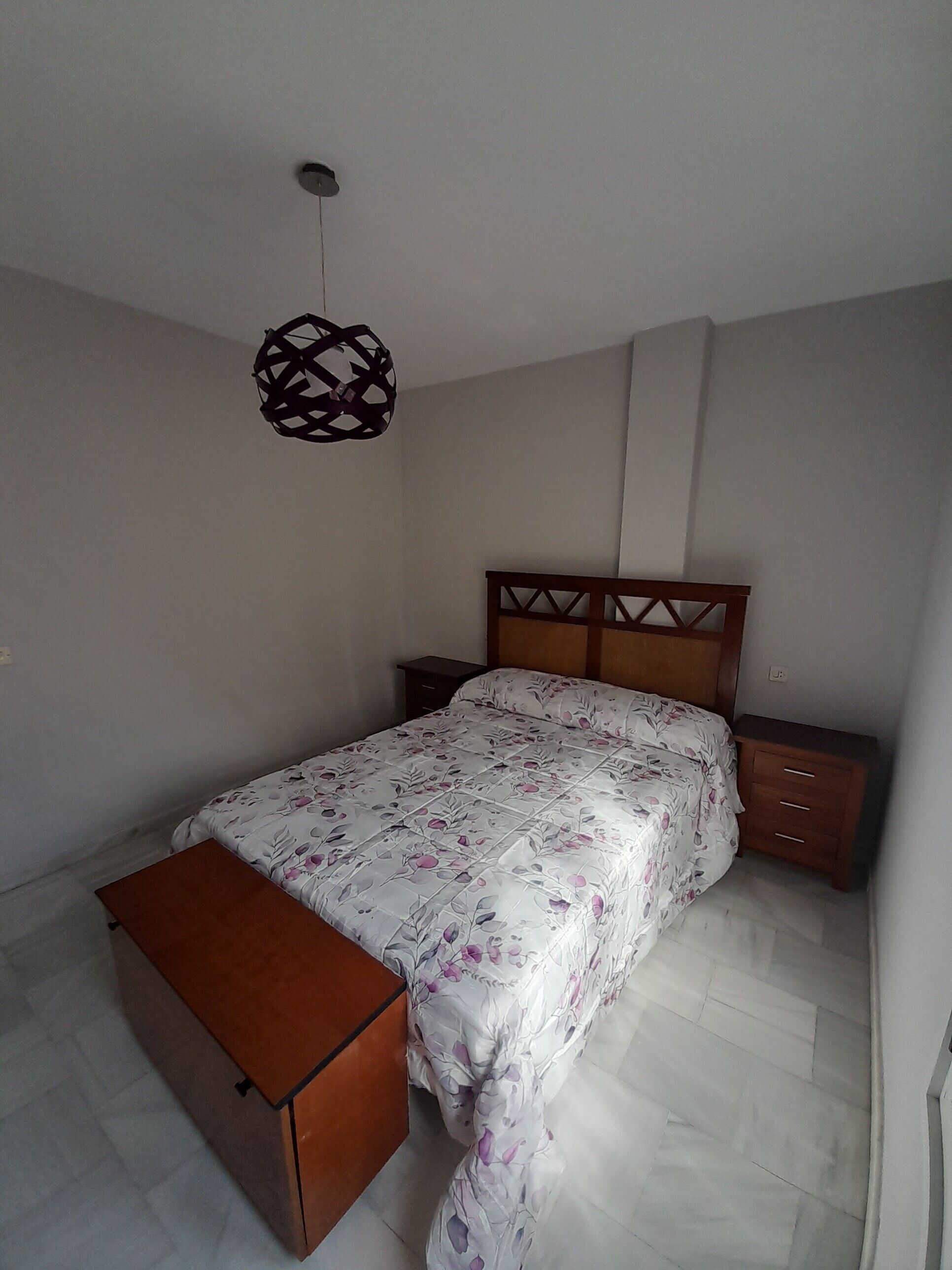 Een slaapkamer met een bed, een houten nachtkastje, een nachttafel, een hanglamp en een klein houten kastje.