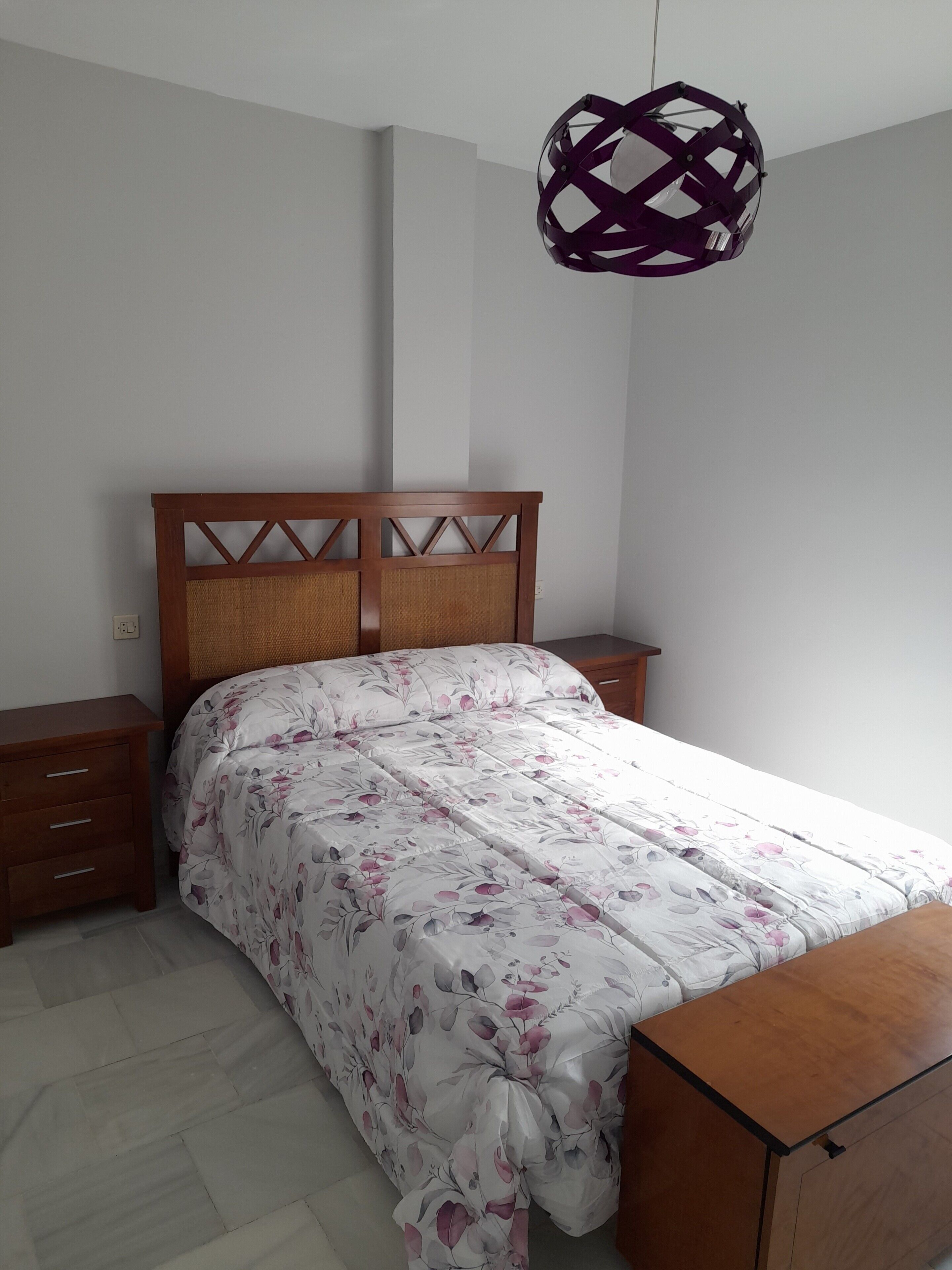 A bedroom with a wooden headboard, a floral bedspread, a wooden nightstand, a wooden dresser, and a hanging lamp.