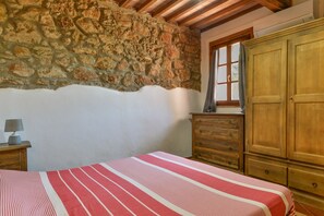 Ein Schlafzimmer mit Steinwand, Holzbalkendecke, einem Bett mit gestreifter Bettdecke, einem hölzernen Schminktisch und einem Fenster mit Vorhängen.