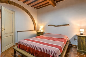 Un dormitorio con cama, un banco de madera, una mesita de noche con lámpara, un estante en la pared y una puerta.