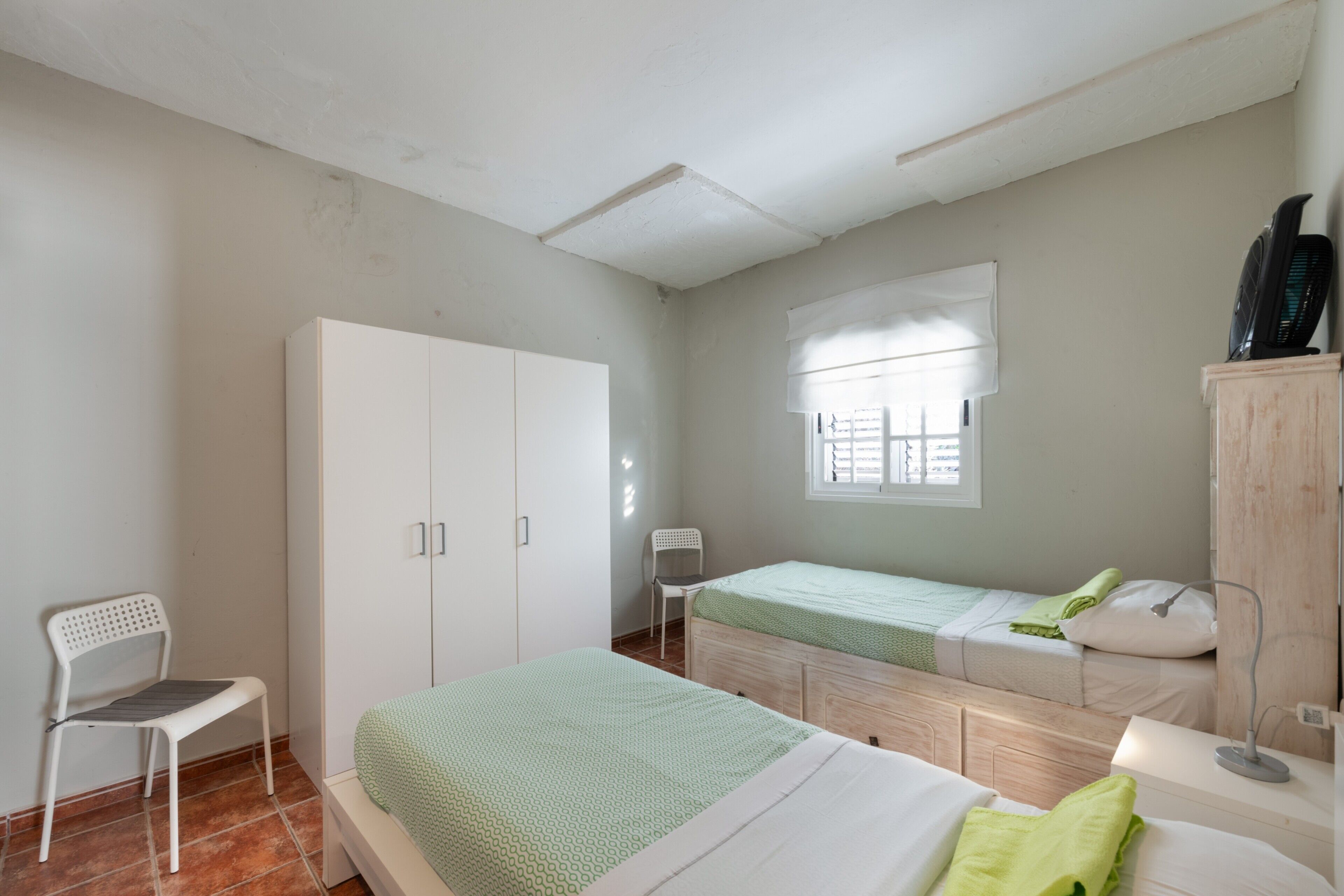 A bedroom with two beds, a wardrobe, a chair, a window with blinds, and a desk with a lamp.