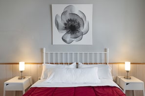 Un lit avec une literie blanche et une couverture rouge, flanqué de deux tables de chevet avec des lampes, un chevet blanc et une œuvre d’art représentant une fleur, accrochée au mur.
