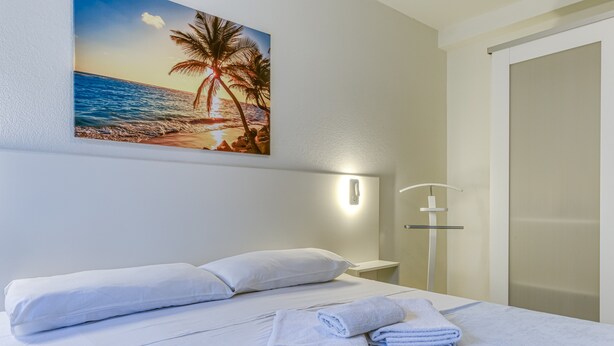 A hotel room with a bed, white linens, a wall-mounted painting of a beach, a nightstand, and a door.