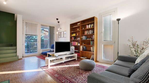Ein Wohnzimmer mit Flachbildfernseher, Bücherregal, grauem Sofa und einem roten Teppich mit Muster.