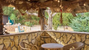 A stone wall with wicker chairs and a table, a rustic wooden ceiling, and a view of a garden with trees and a stone structure. - Villa 'Santa Ana' with Private Pool, Wi-Fi and Air Conditioning (La Carlota)