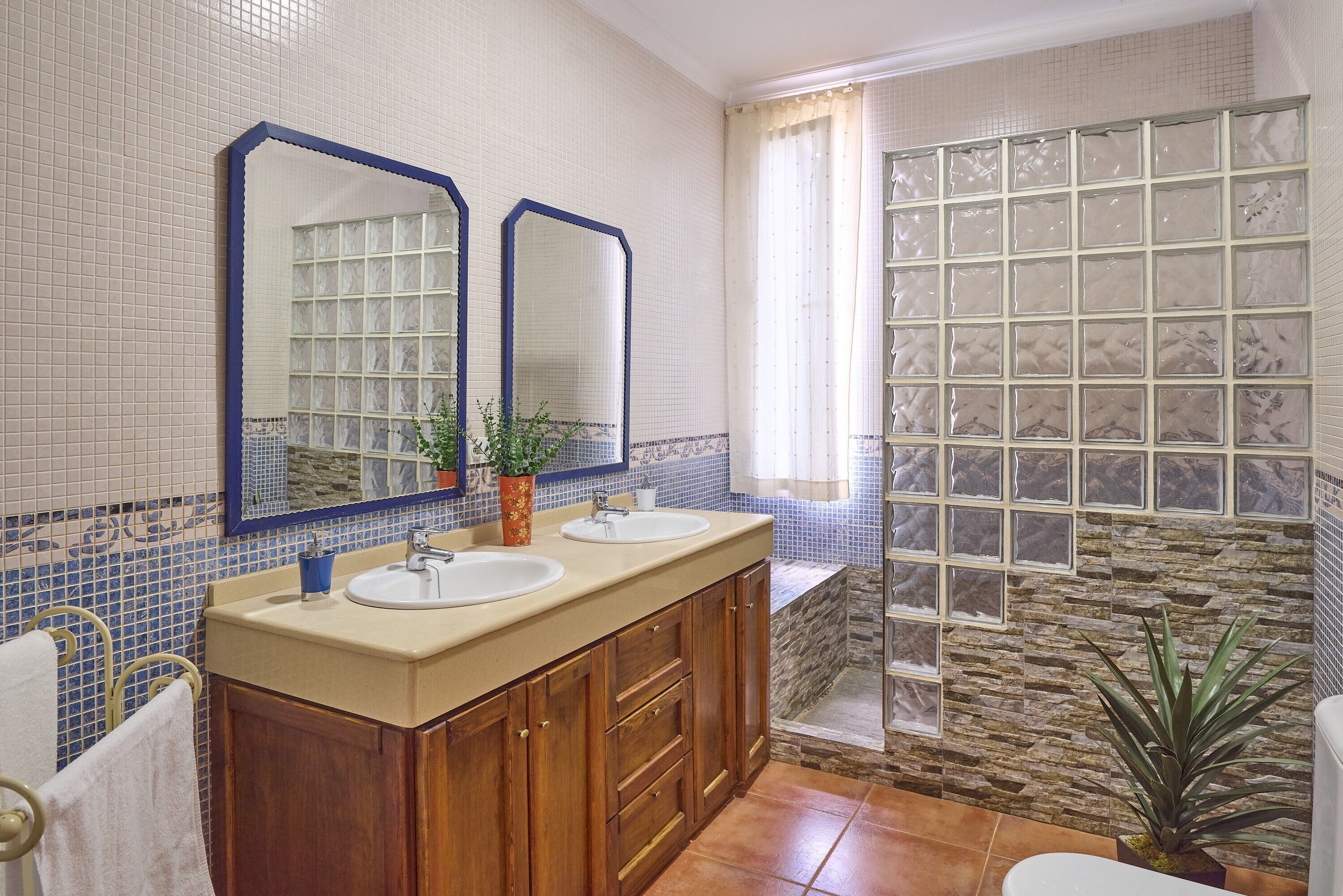 A bathroom with two sinks, wooden cabinets, a mirror, a potted plant, and a shower area with a stone wall.